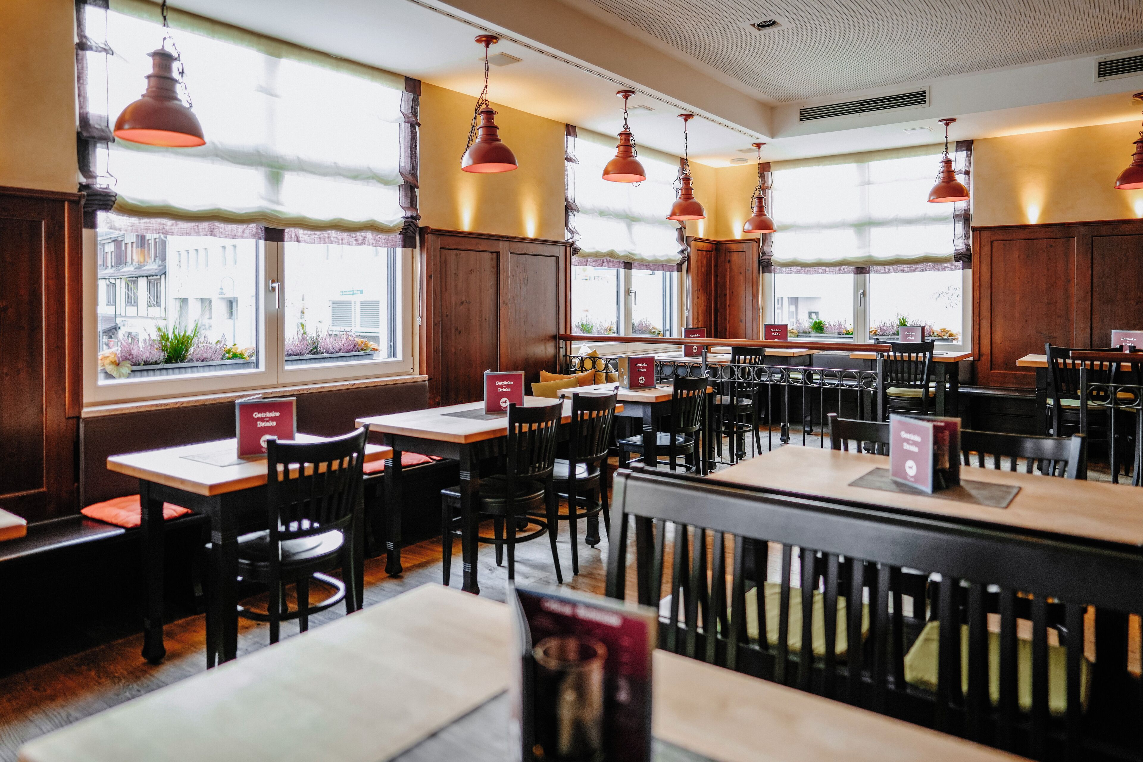 A restaurant interior with wooden tables and chairs, pendant lighting, and large windows with views of buildings and plants outside.