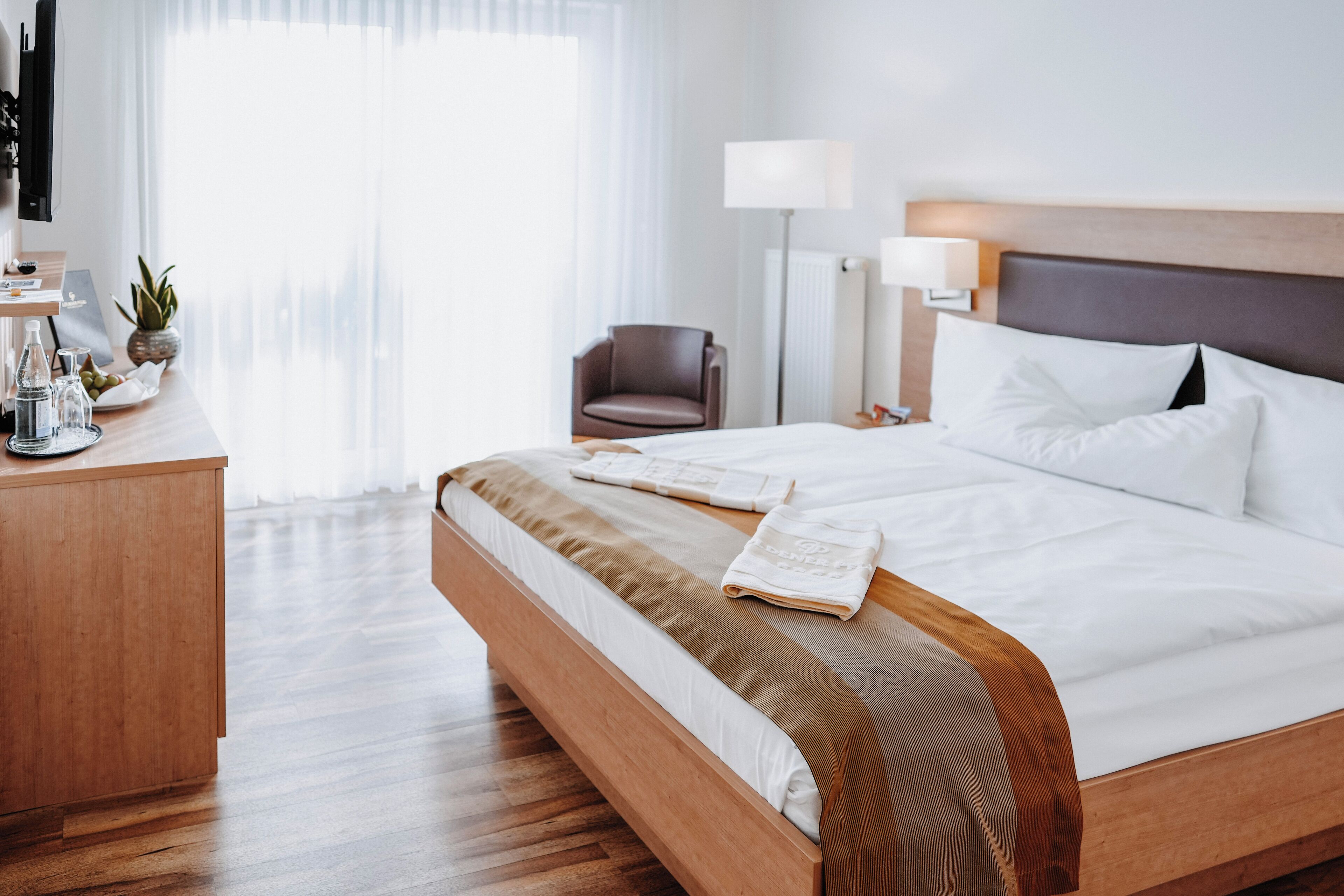 Habitación de hotel con una cama de madera, un escritorio con lámpara, una silla y vistas a la ciudad a través de amplias ventanas.