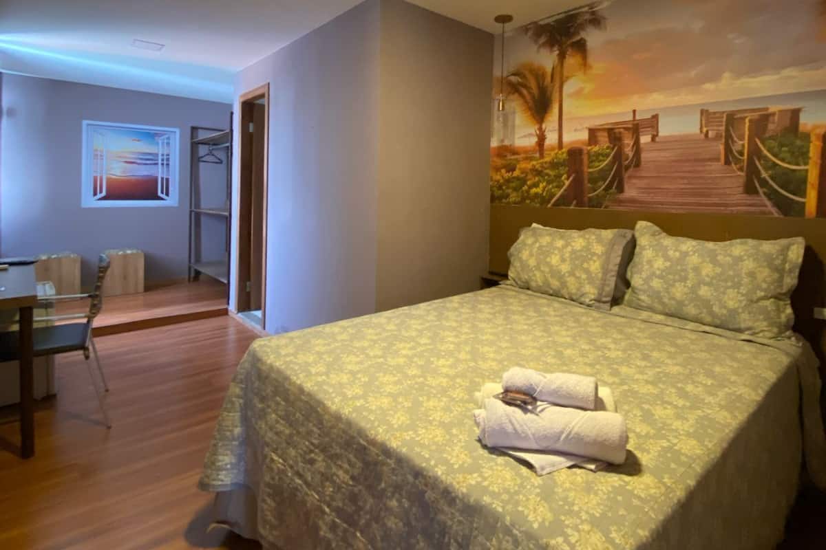 A bedroom with a bed, a desk, and a chair. There is a painting on the wall depicting a beach scene.