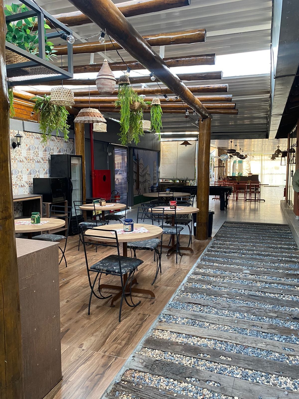 Interior de un restaurante con mesas de madera, plantas colgantes y una pared de azulejos.
