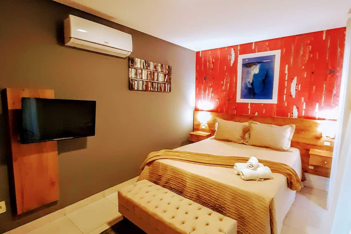 A modern hotel room with a flat-screen TV, a large bed with a matching bench, a red accent wall, and a small bedside table with a lamp.