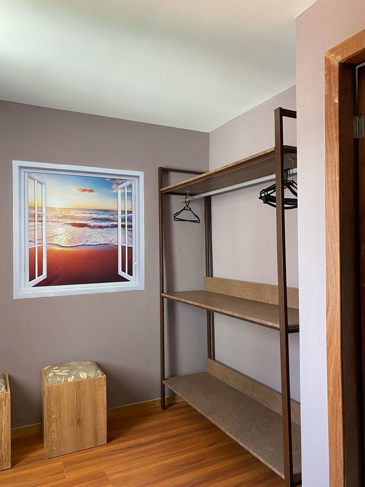 Una habitación con literas, un taburete de madera, un perchero y una ventana con vistas a un atardecer sobre el océano.