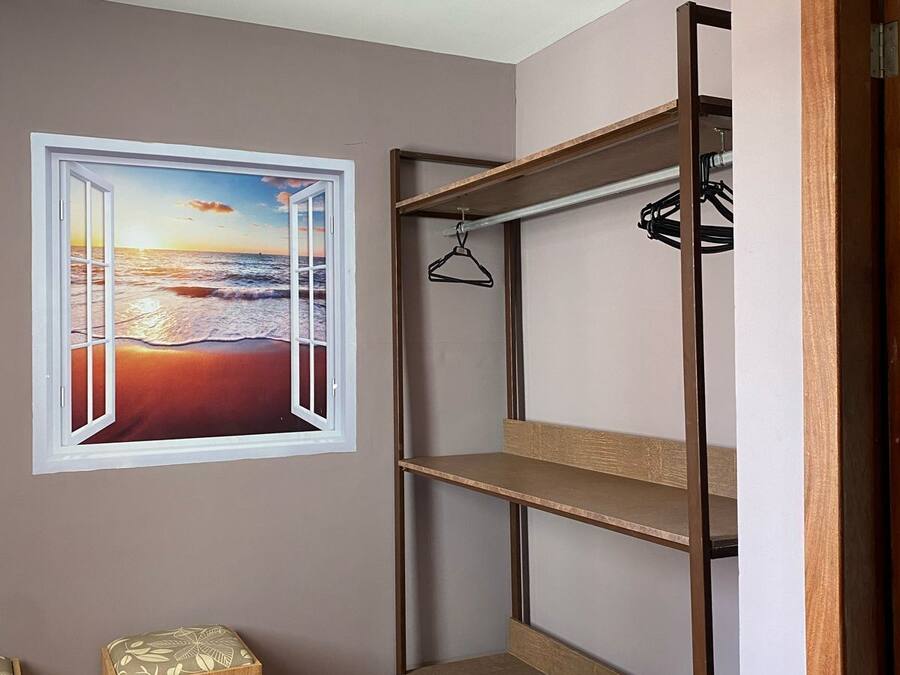 A room with a bunk bed, a wooden stool, a hanging clothes rack, and a window with a view of a sunset over the ocean.