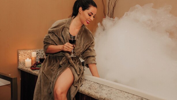 A person in a robe sitting by a steam shower, holding a glass. - Serra Negra Pousada Spa by Easy Hoteis (Guarapari)