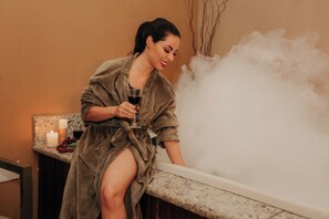 A person in a robe sitting by a steam shower, holding a glass.