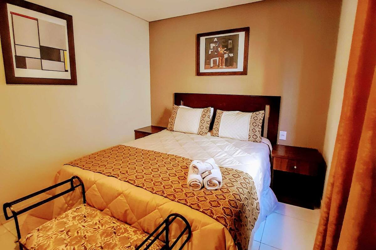 A hotel room with a bed, a bedside table, a chair, and a small table with towels.