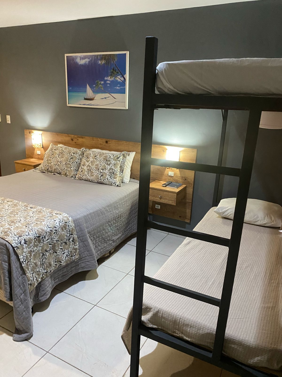A bedroom with a bunk bed, a single bed, a nightstand with a lamp, and a painting on the wall.