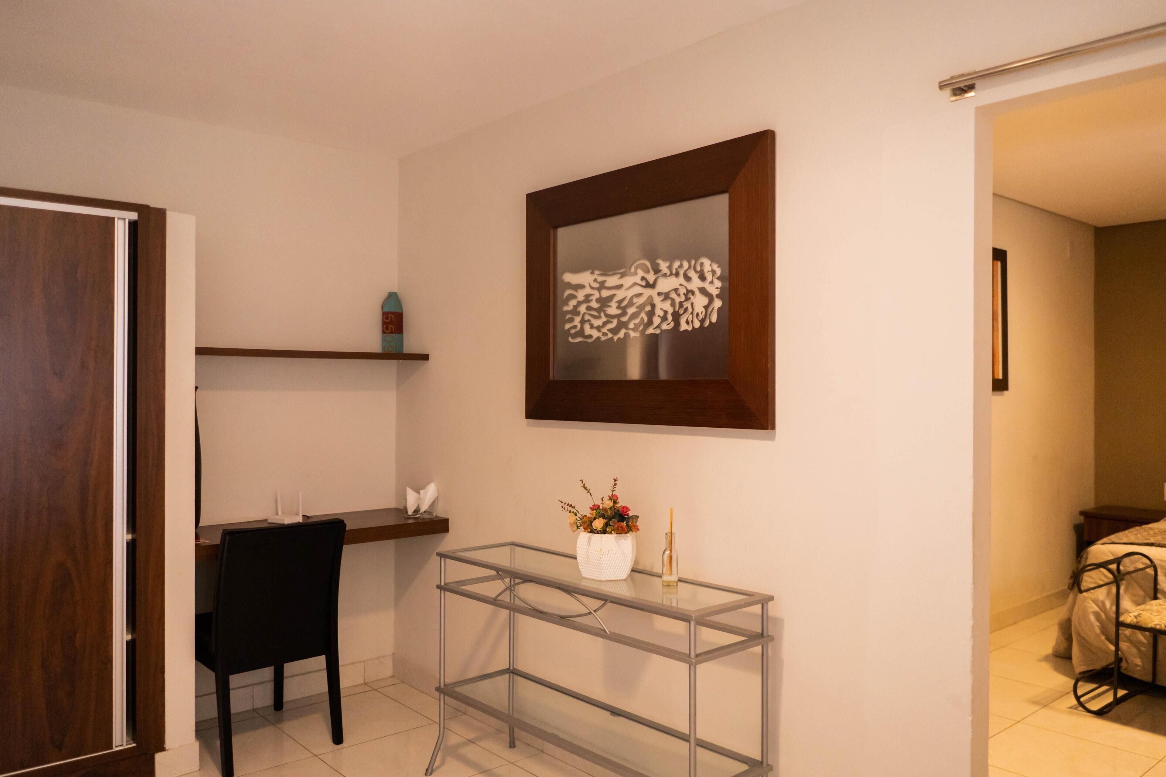A room with a glass table, a chair, a shelf with decorative items, and a framed artwork on the wall.