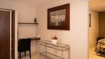 A room with a glass table, a chair, a shelf with decorative items, and a framed artwork on the wall.