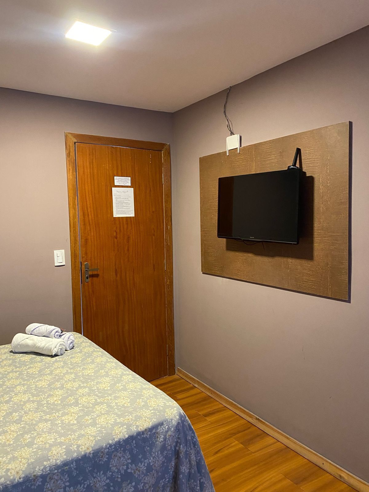 Quarto de hotel com cama, porta de madeira, televisão montada e luminária.