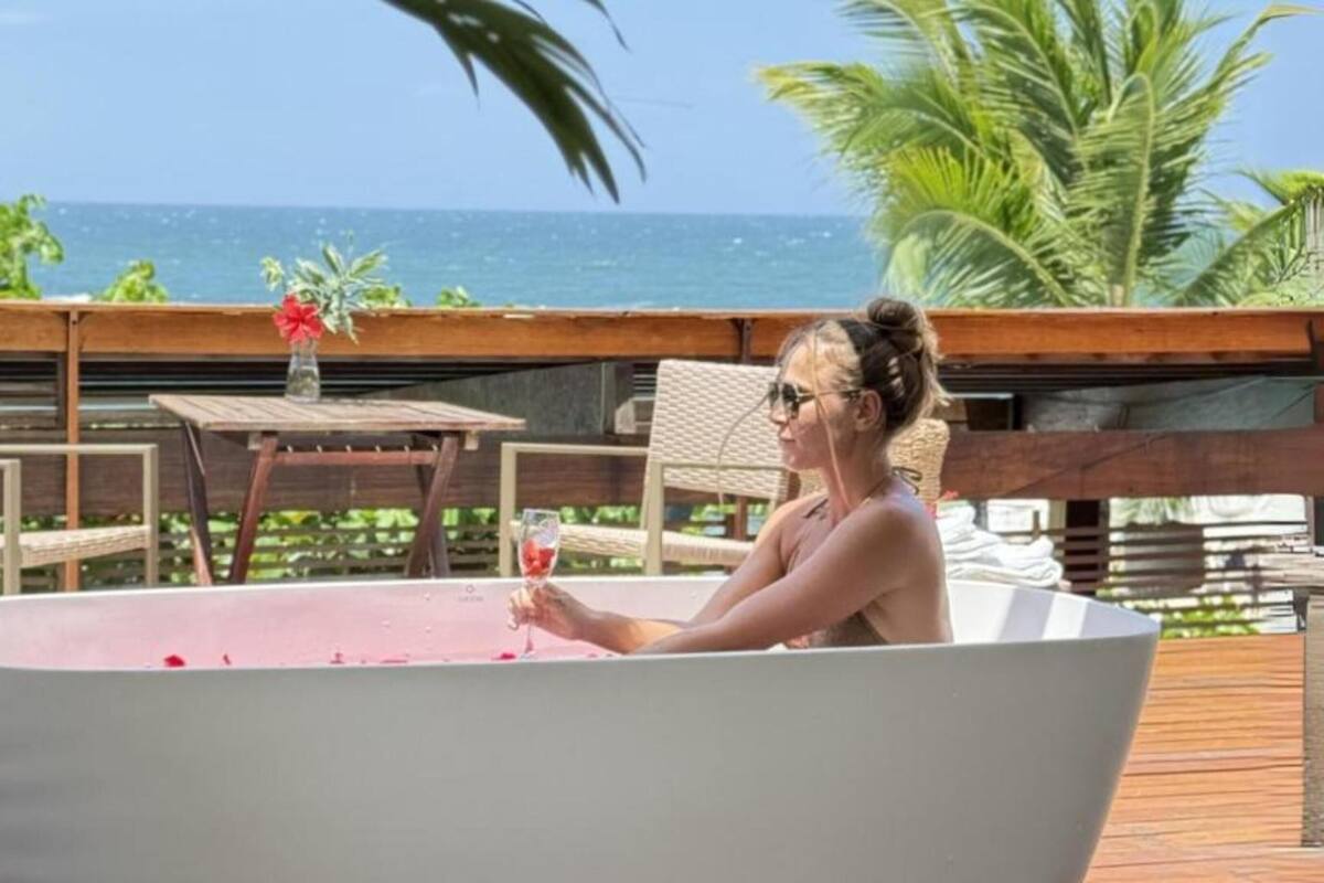 A woman in a large bathtub on a wooden deck with a table and chairs, overlooking the ocean.