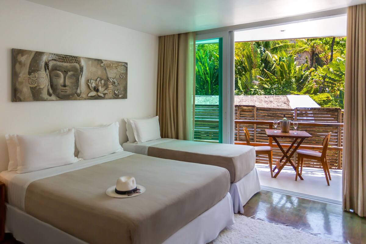A hotel room with two beds, a painting on the wall, a dining area with a table and chairs, and a view of greenery outside.