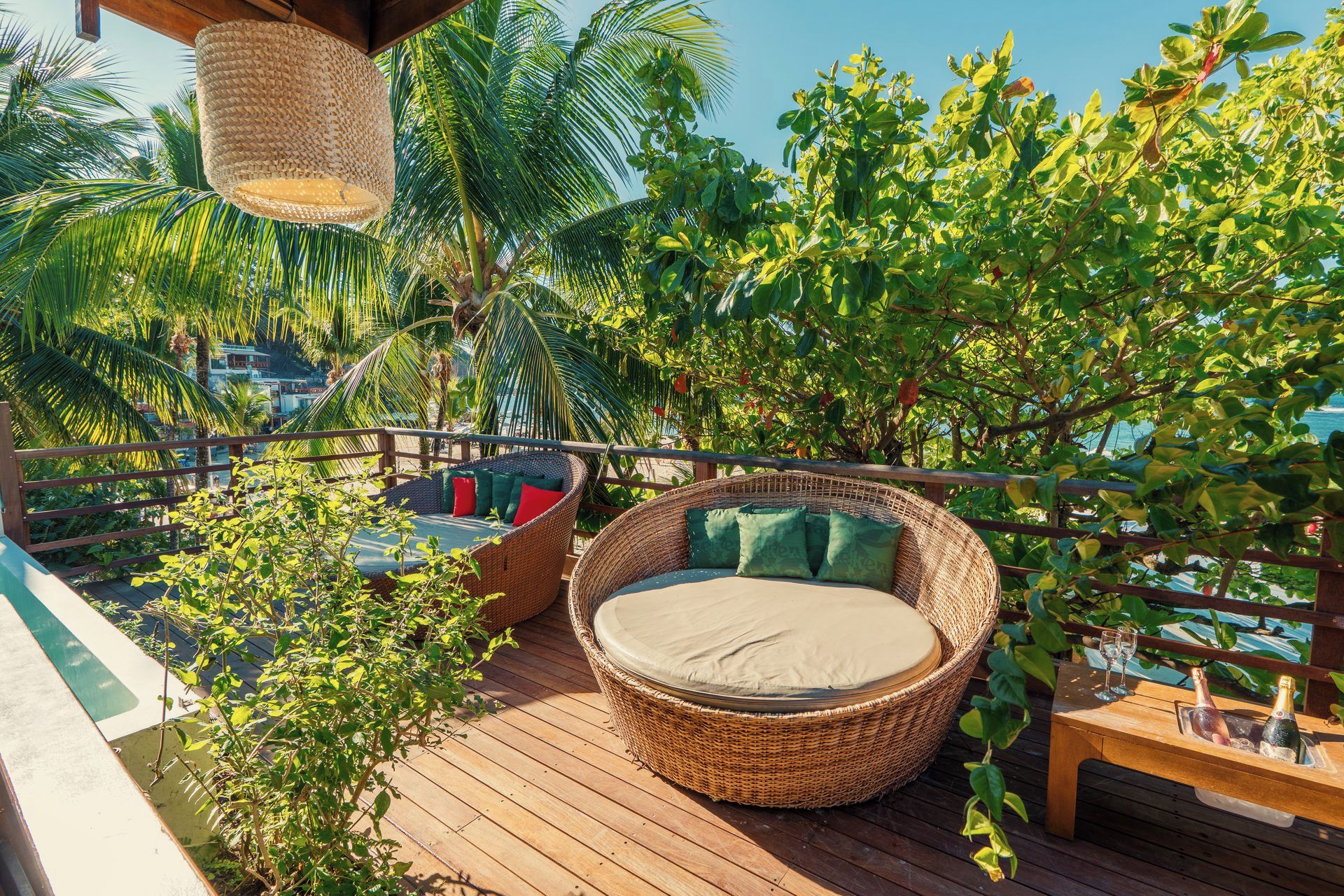 Une terrasse en bois agrémentée de meubles en osier, notamment un fauteuil rond et un canapé avec des coussins, le tout entouré d’une végétation luxuriante et offrant une vue sur la mer.