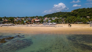 Una playa con aguas transparentes y poco profundas, hileras de sombrillas y un fondo de edificios coloridos y abundante vegetación.