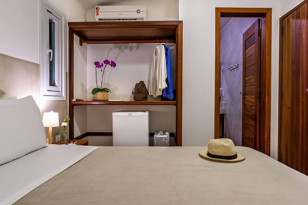 A bedroom with a bed, a bedside table with a lamp, a small shelf with a plant, a suitcase, and a hat on the bed.