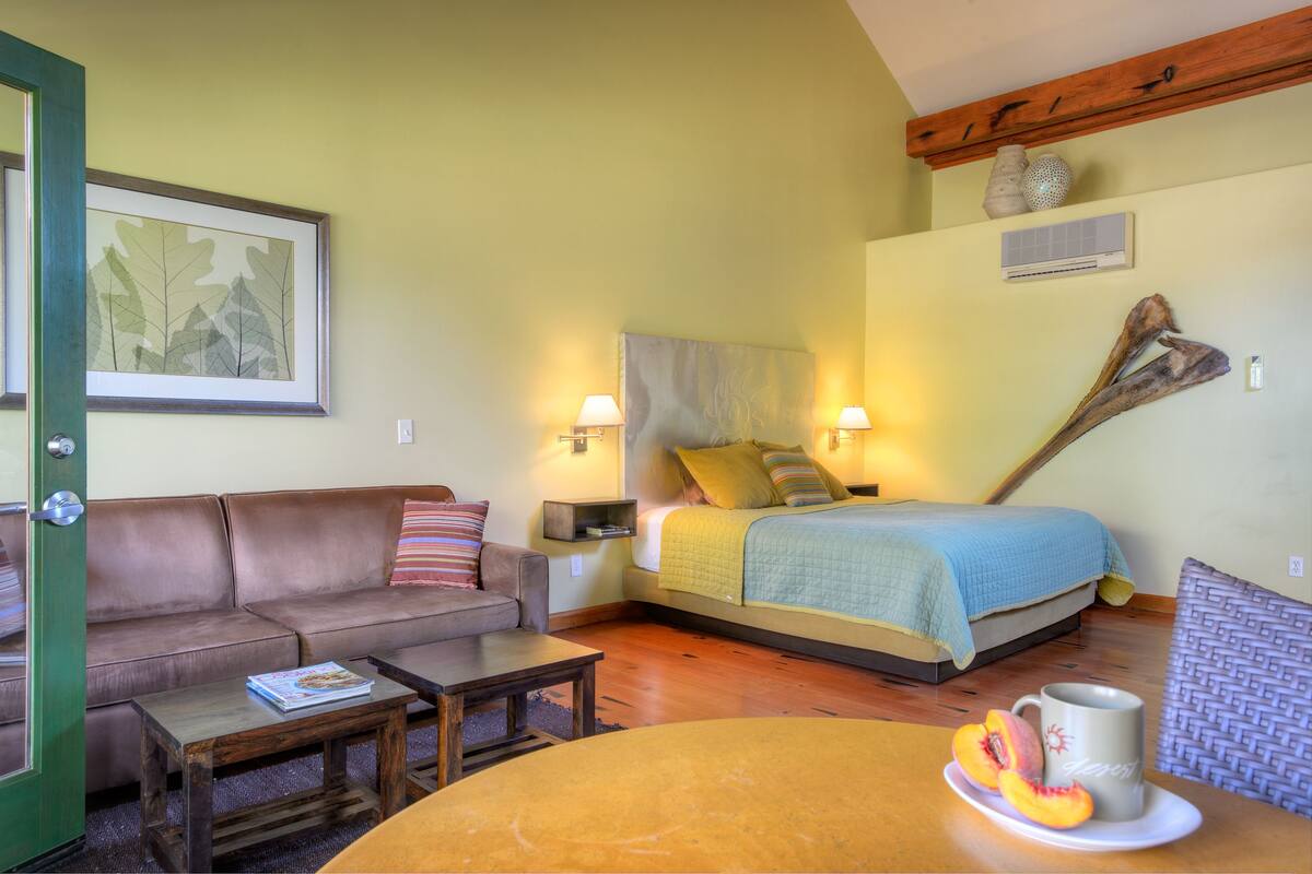 A bedroom with a bed, a sofa, a coffee table, and a dining table with fruit and a mug.