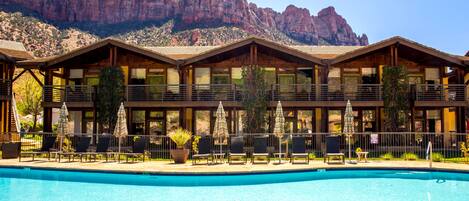 Un hotel con piscina, mobiliario de exterior y montañas al fondo.