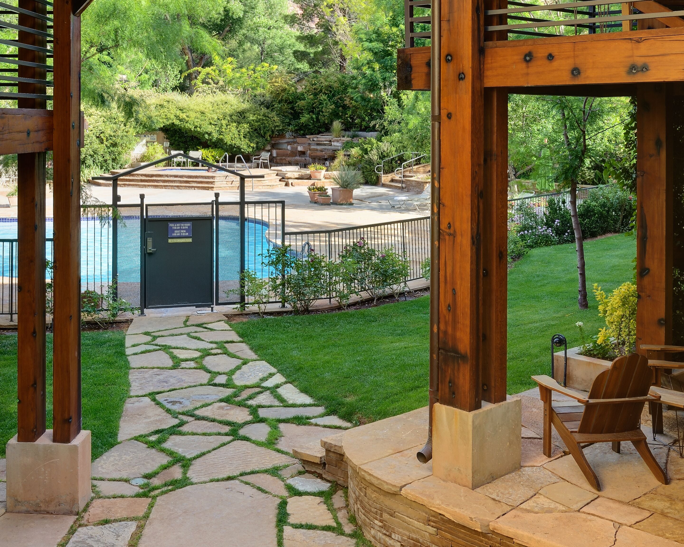 A backyard with a stone pathway leading to a pool area, surrounded by greenery and a pergola structure.