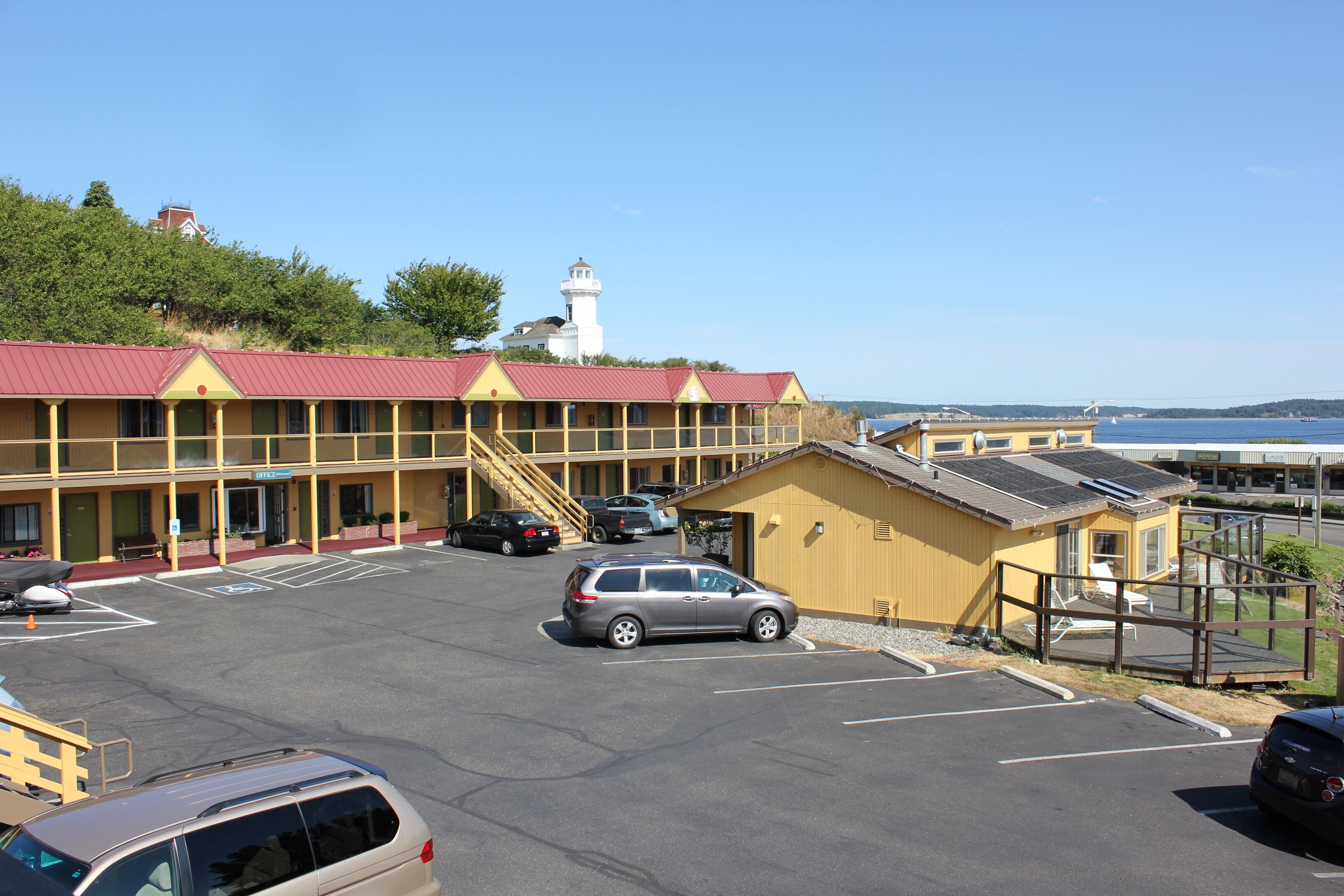 A motel with a red roof, a parking lot, and a view of a lighthouse in the background.