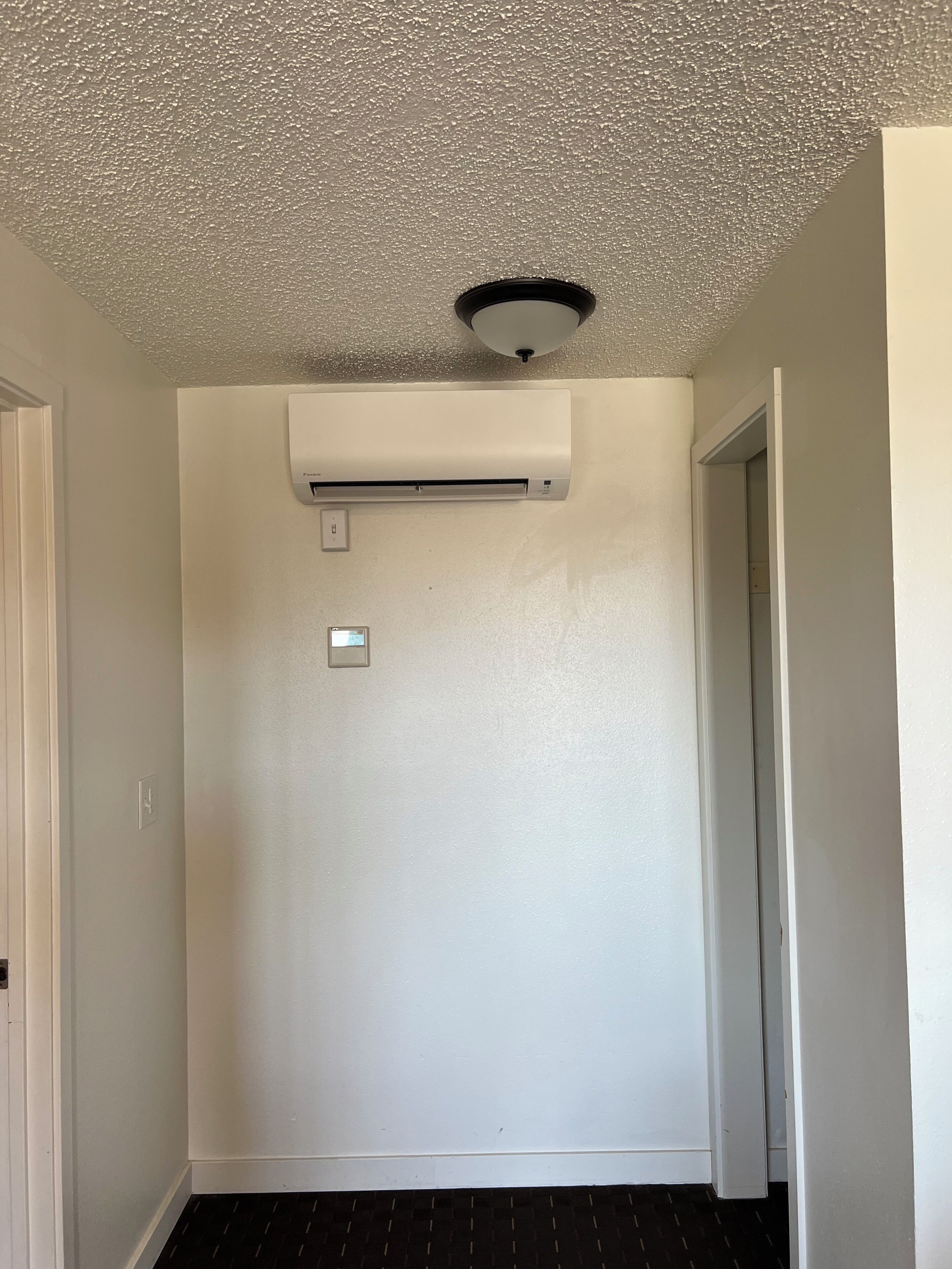 A room with a white air conditioning unit mounted on the wall, a ceiling light fixture, and a door leading to another room.