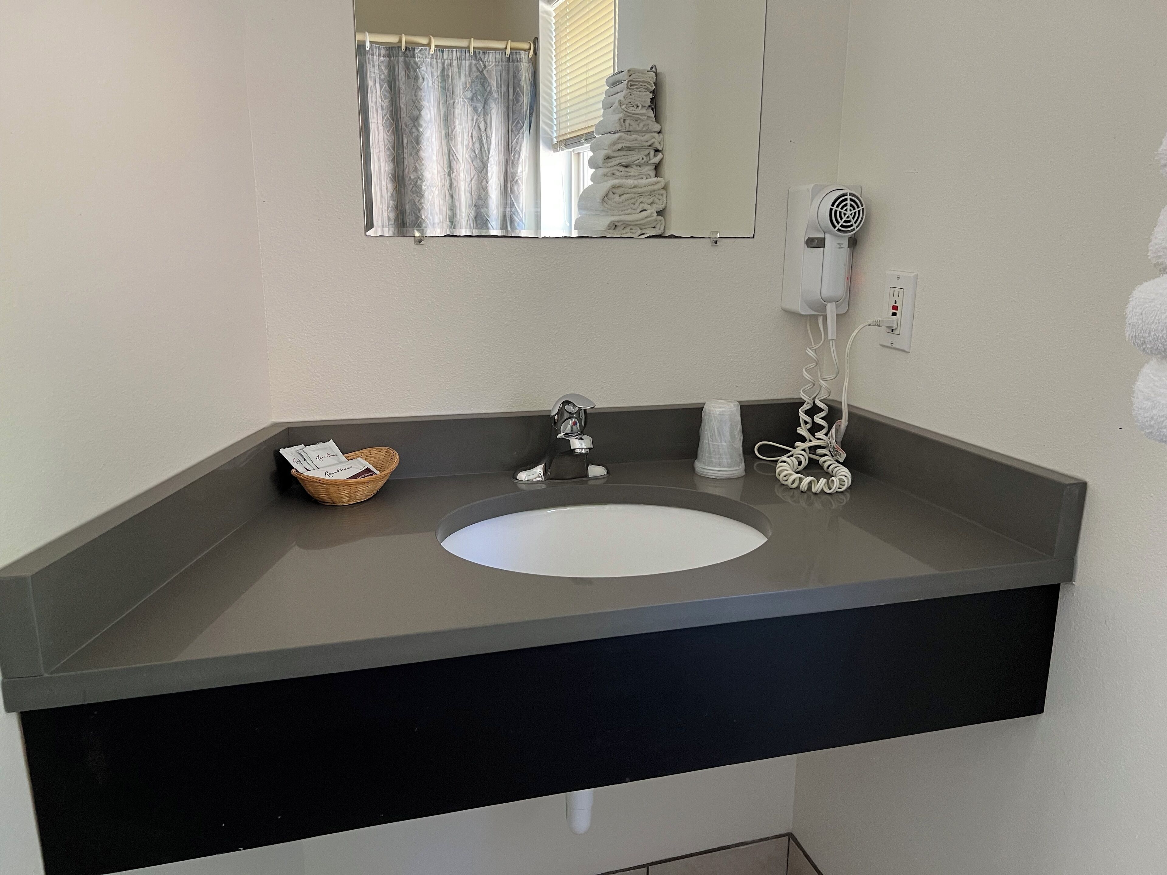 Un baño con un lavamanos blanco, un espejo, un perchero con toallas dobladas, una cesta con artículos de aseo personal y un secador de pelo enchufado.
