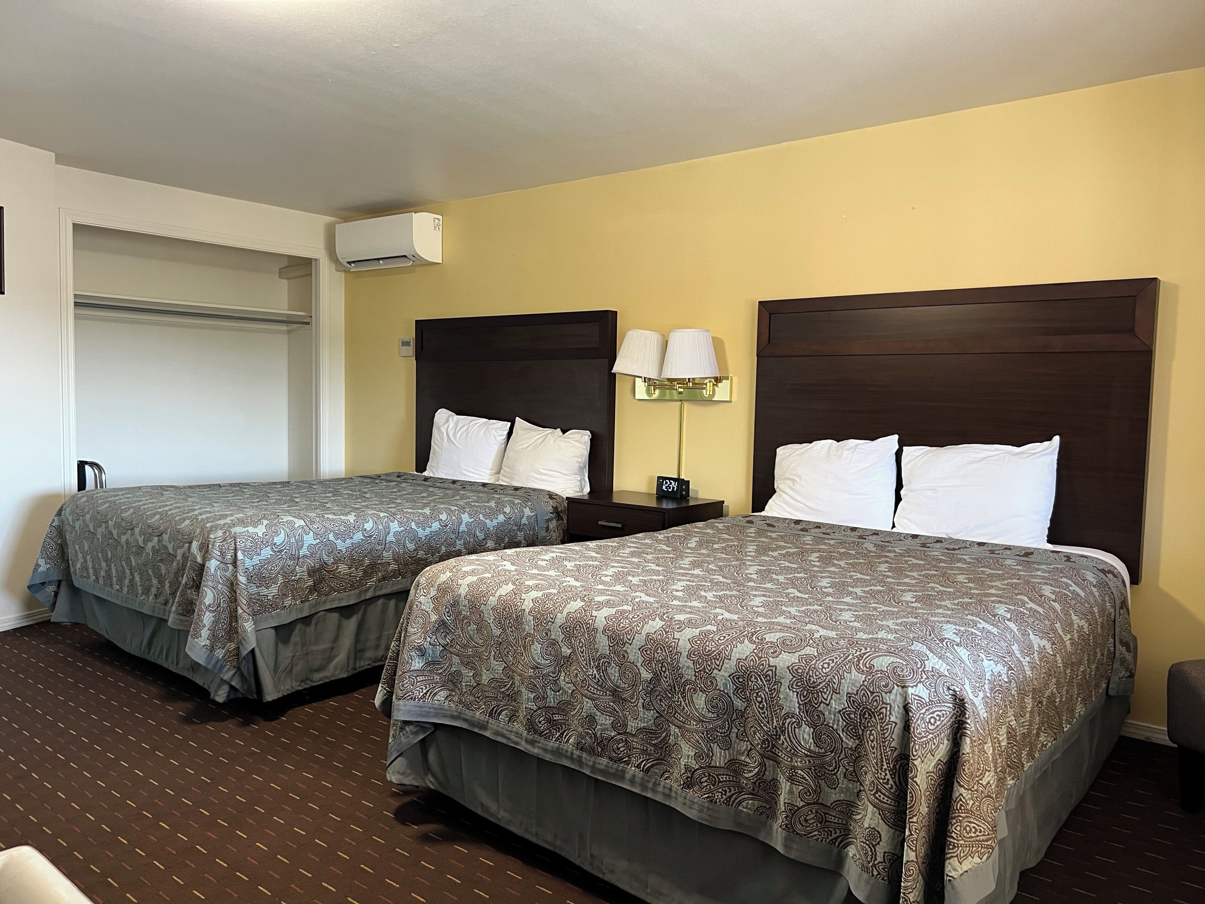 A hotel room with two beds, a nightstand with a lamp, a clock, and a wall-mounted air conditioner.