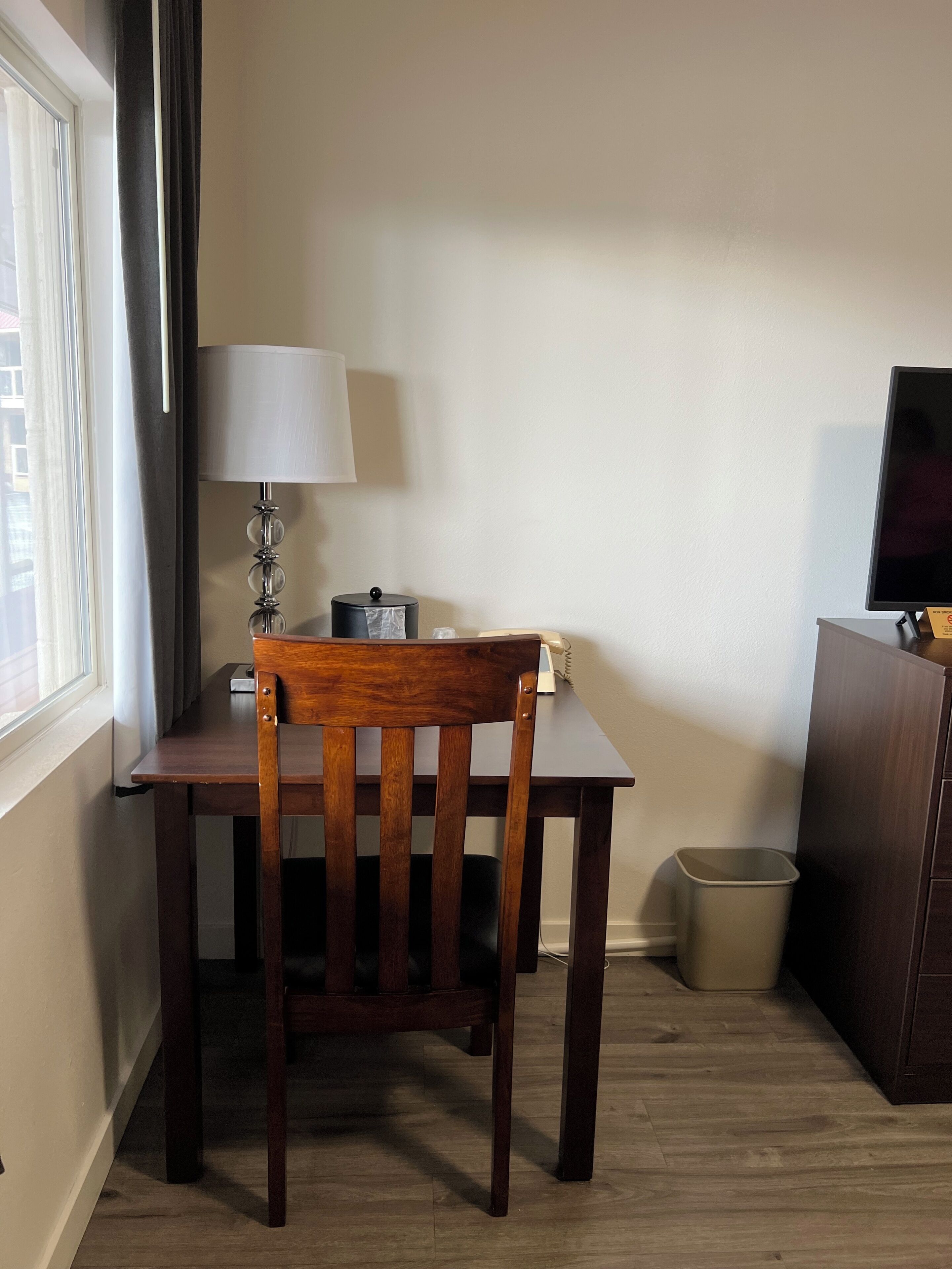 A wooden table with a chair, a lamp, a television, and a trash bin in a room with a window and a white wall.