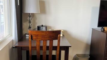 A wooden table with a chair, a lamp, a television, and a trash bin in a room with a window and a white wall.