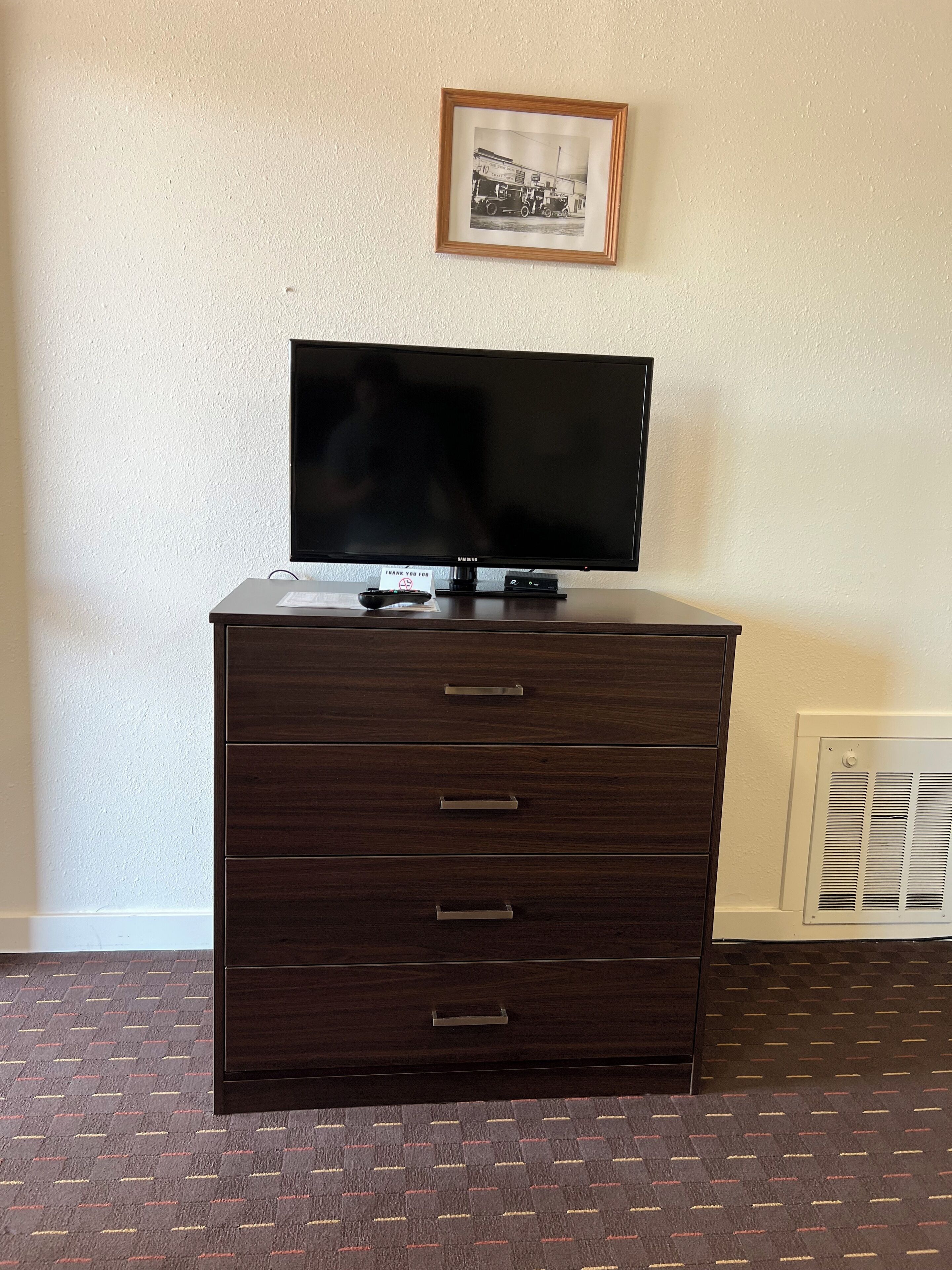 A dark wooden dresser with a flat-screen TV on top, a framed photo on the wall, and a vent on the wall.