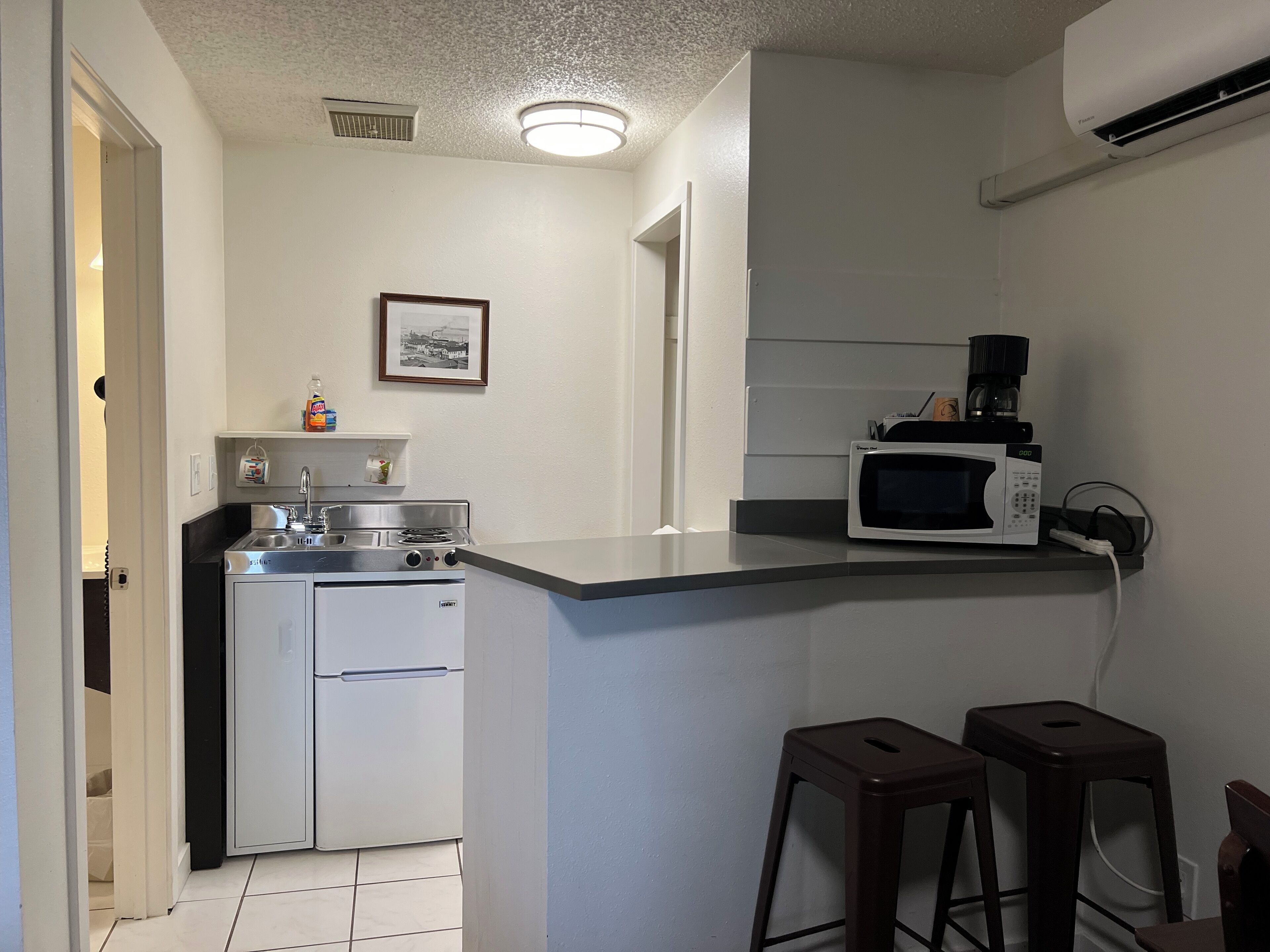 Uma cozinha compacta com fogão, micro-ondas e cafeteira. Há banquetas e um quadro na parede.
