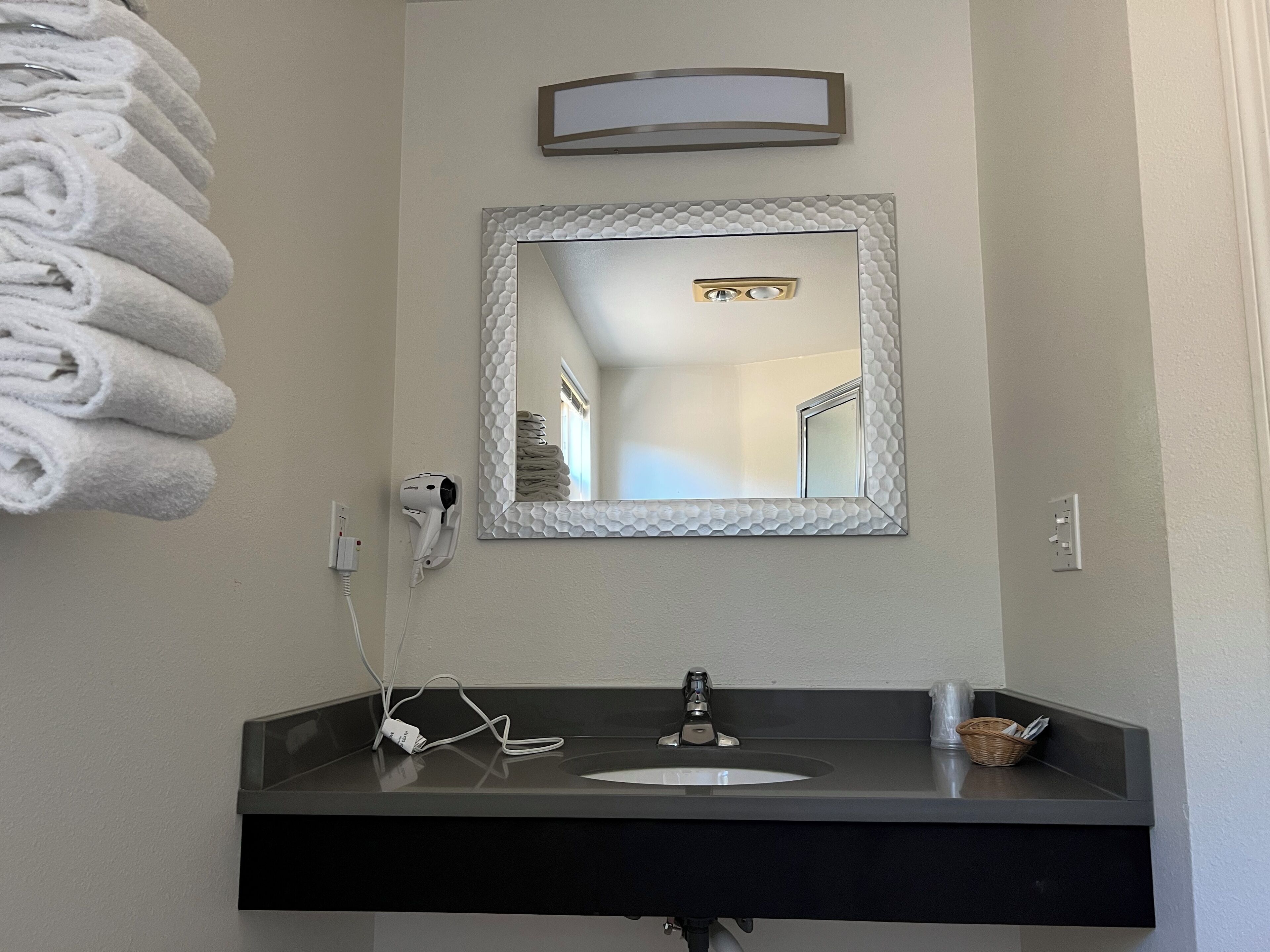 A bathroom with a white towel rack, a hairdryer, a mirror with a textured frame, a sink with a black countertop, and a basket on the countertop.