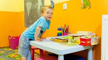 Een kind aan een tafel met leermateriaal, een schoolbord met tekeningen en een dinosaurusdecoratie aan de muur.
