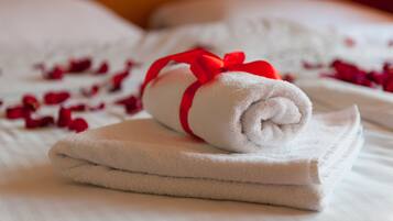 A neatly folded white towel with a red ribbon on a bed with white sheets and scattered rose petals.