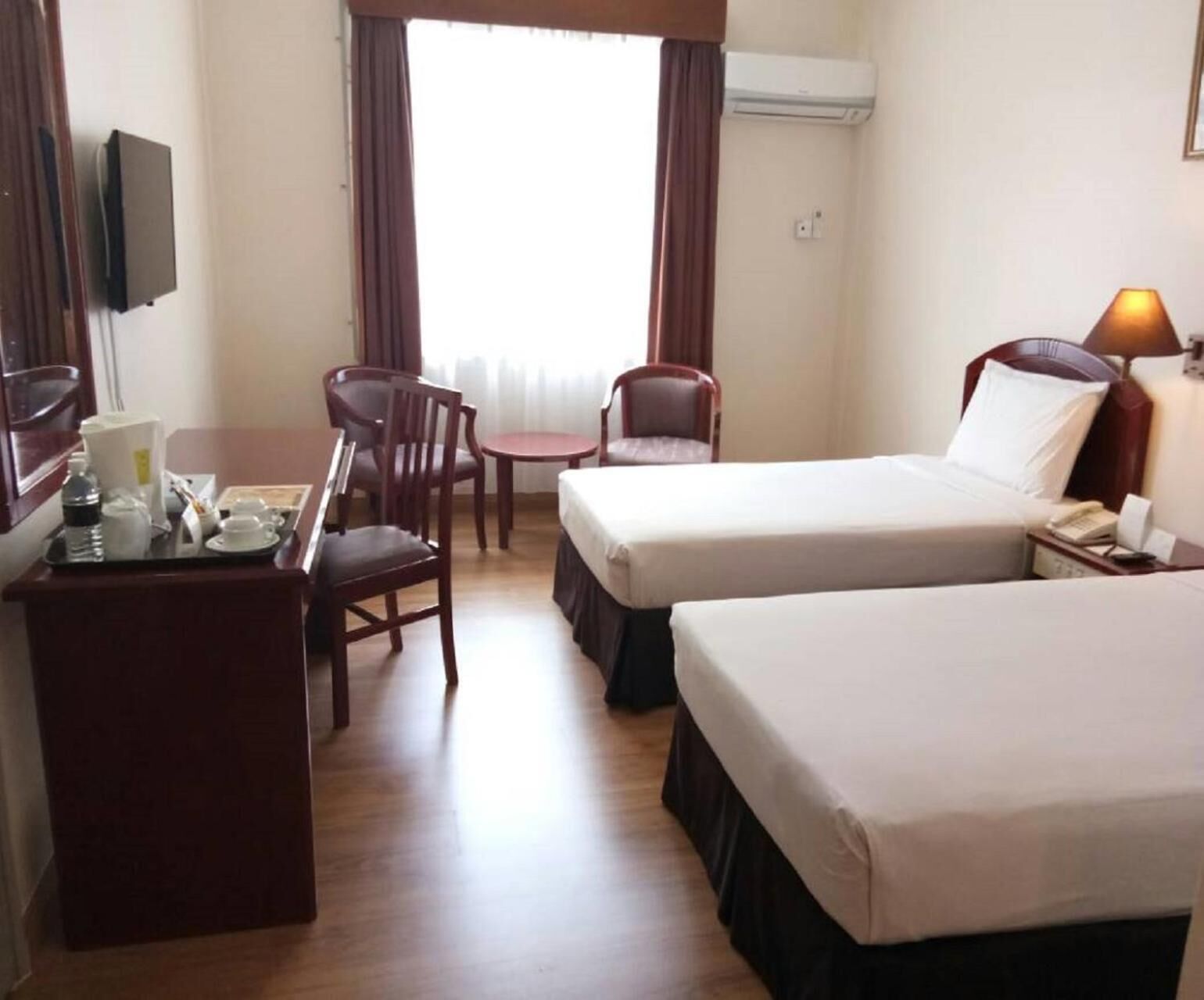 A hotel room with two beds, a desk with a chair, a TV, and an air conditioning unit.