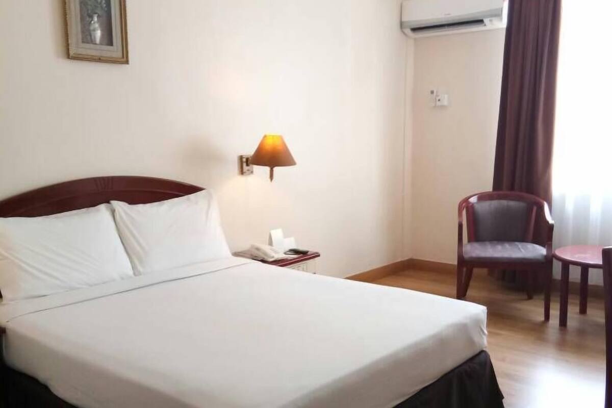 A hotel room with a large bed, a bedside table with a lamp, a chair, an air conditioning unit, and a framed picture on the wall.