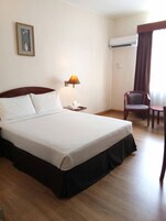 Una habitación de hotel con una cama grande, una mesita de noche con lámpara, una silla, un aire acondicionado y un cuadro en la pared.