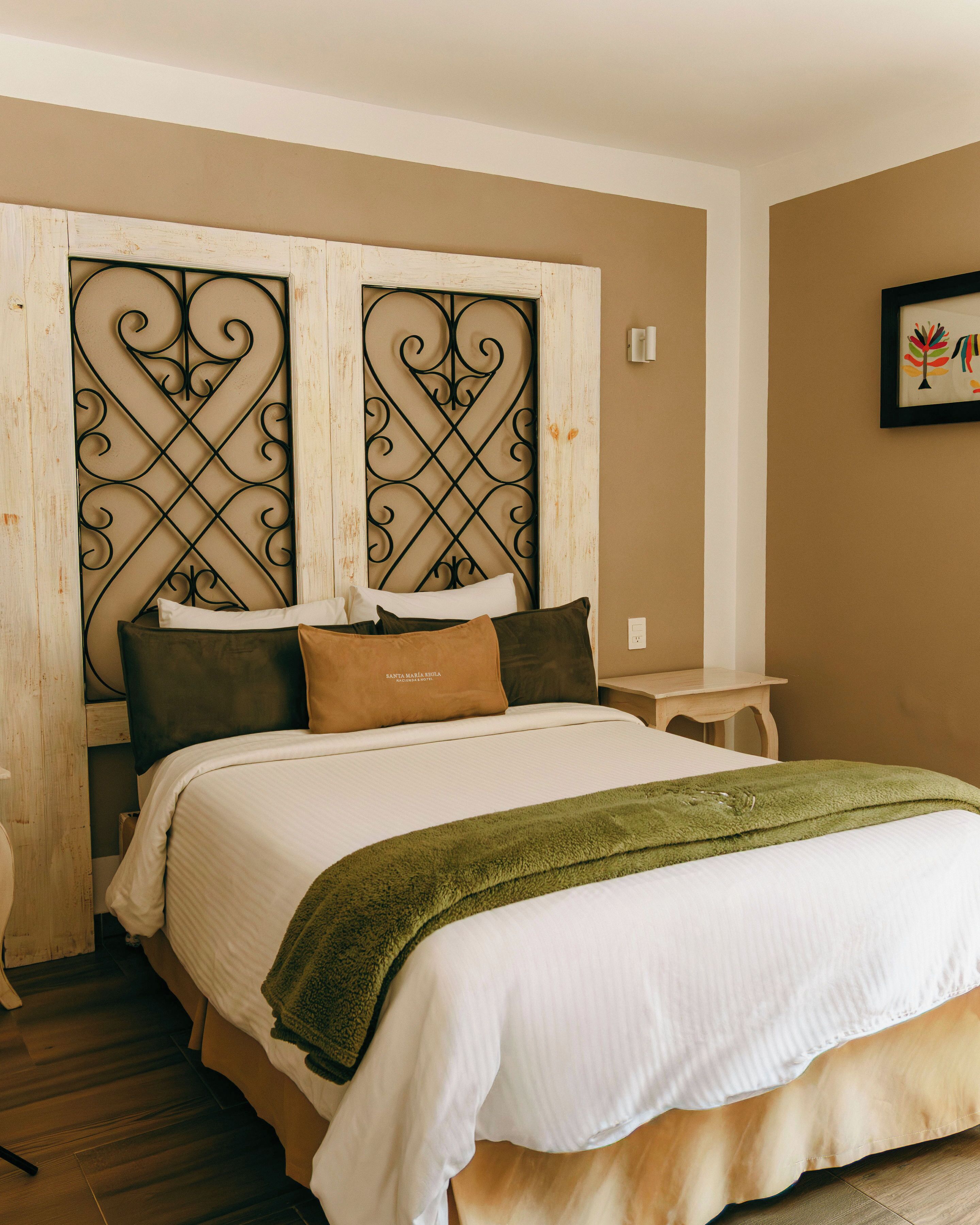 Un lit bien fait, recouvert d’une couverture verte, avec une tête de lit en bois ornée d’un motif métallique décoratif et une petite table de chevet.