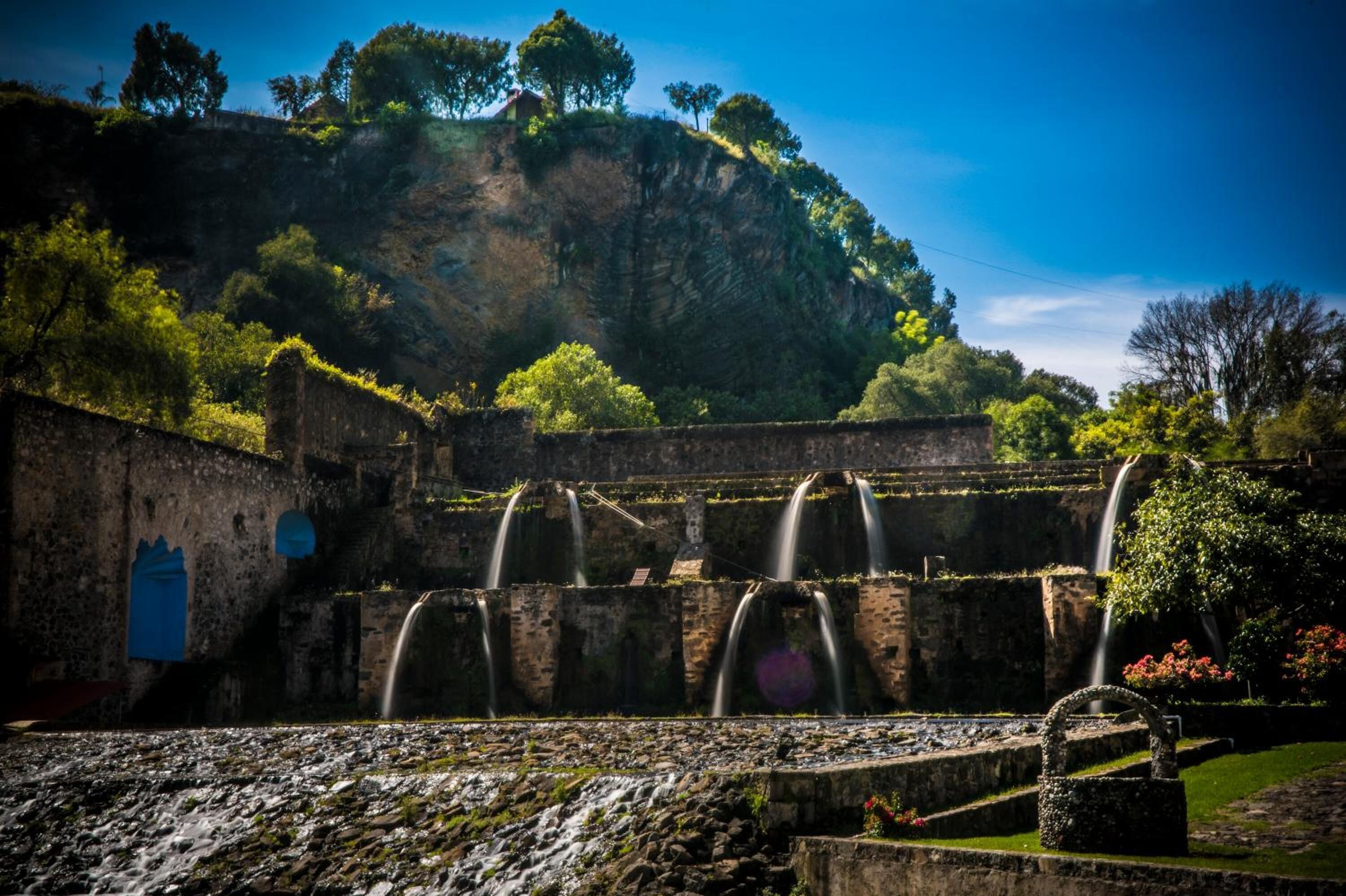 An ancient stone structure with multiple water channels and a lush green hillside in the background.