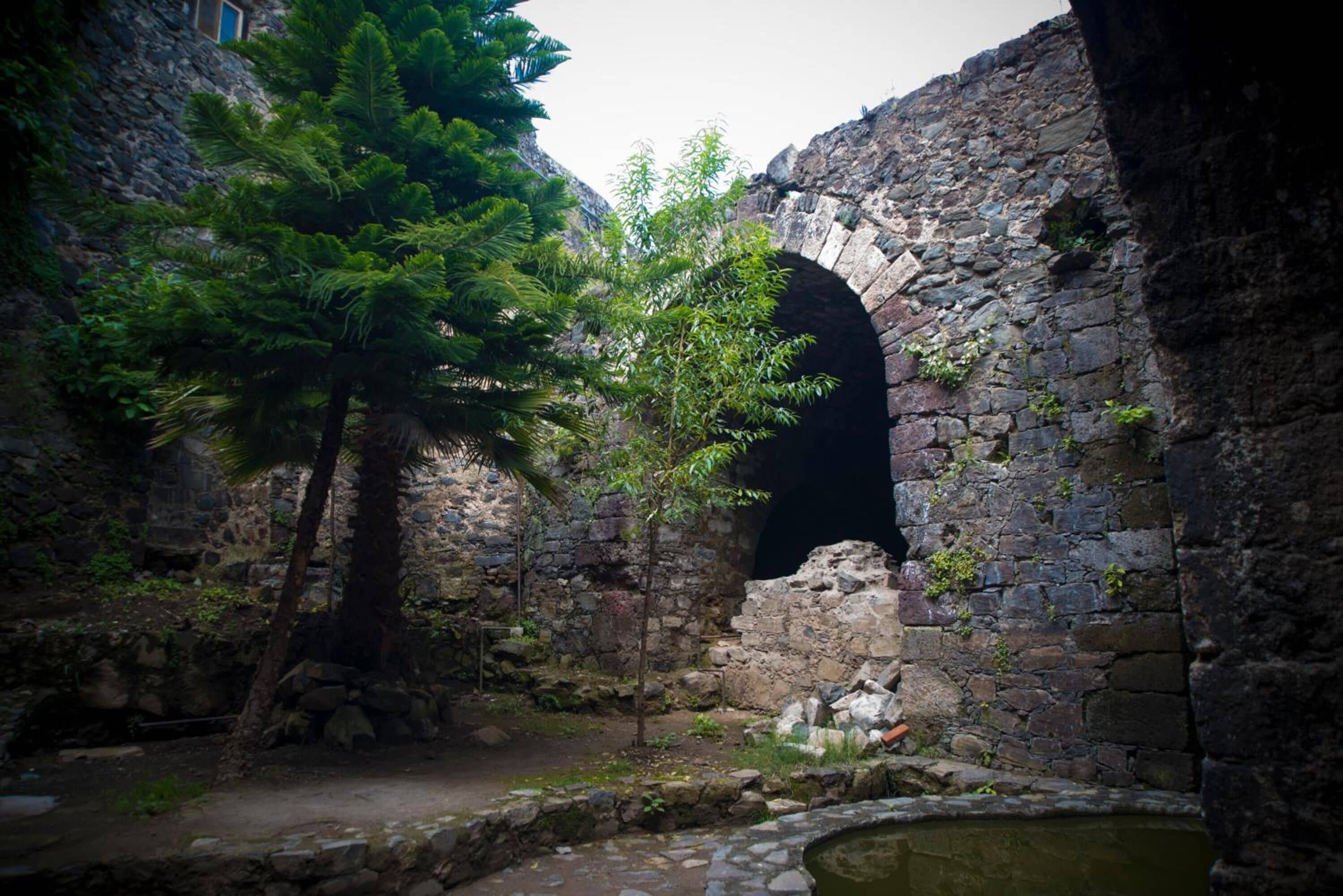 An ancient stone structure with a narrow passageway, a small water feature, and lush greenery.