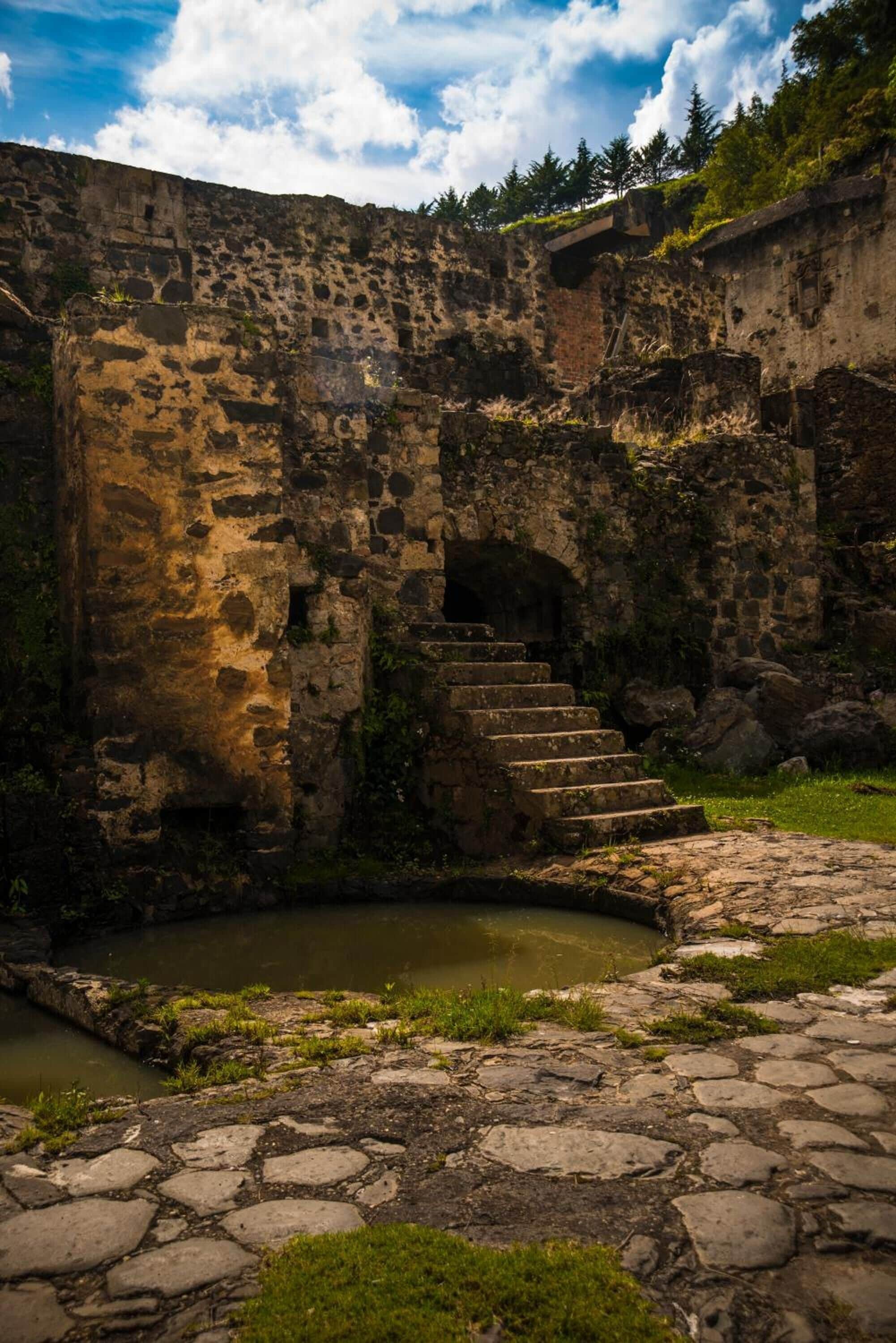 An ancient stone structure with a staircase leading to an arched entrance, surrounded by a small pond and stone pathway.