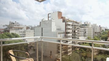 A balcony with wicker chairs, overlooking a cityscape with multiple buildings and trees.
