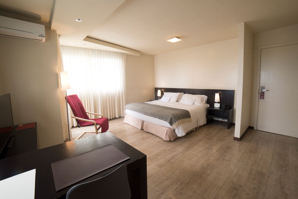 A hotel room with a bed, a desk, a chair, a nightstand, and a window with curtains.