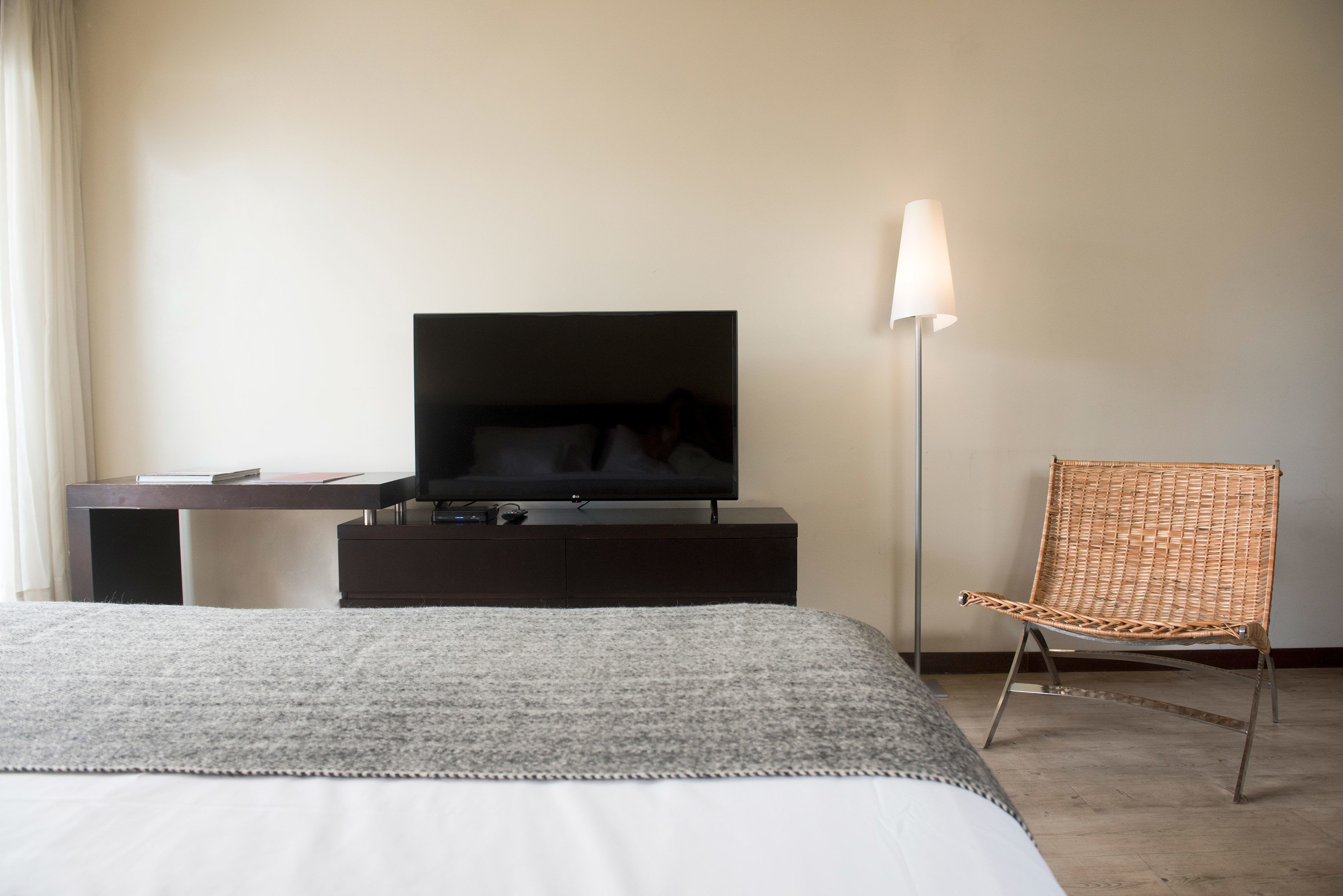 Habitació d'hotel amb un llit, una televisió sobre un suport de fusta fosca, una cadira de vimetla i una làmpada de peu.