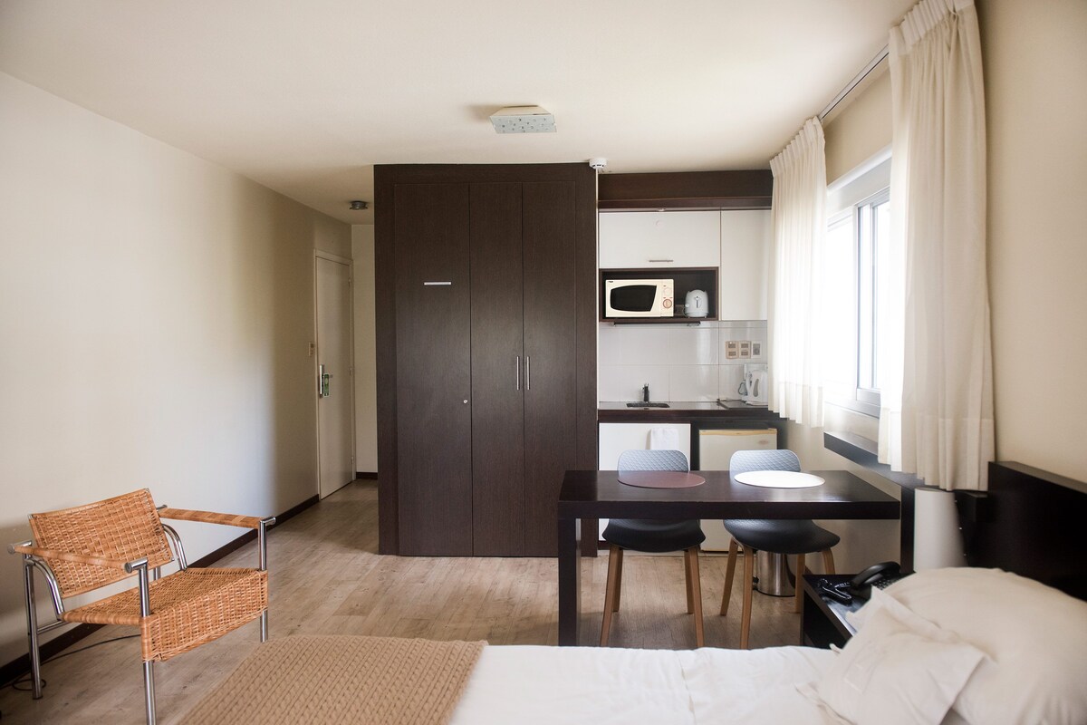 A hotel room with a bed, a dining table with chairs, a microwave, a small kitchen area, and a window with curtains.