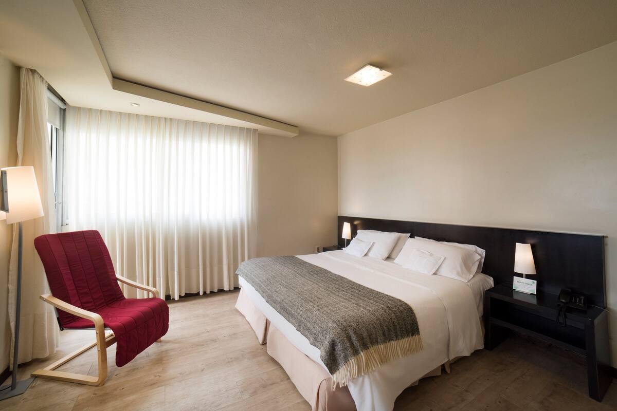 A hotel room with a bed, a red armchair, a bedside table with a lamp, and a window with curtains.