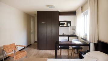 A hotel room with a bed, a dining table with chairs, a microwave, a small kitchen area, and a window with curtains.