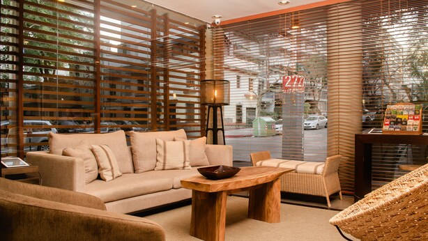 A modern interior with a sofa, wooden coffee table, wicker chairs, and a large window with blinds. - 27 Suites Apart Hotel (Montevideo)