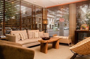 A modern interior with a sofa, wooden coffee table, wicker chairs, and a large window with blinds. - 27 Suites Apart Hotel (Montevideo)