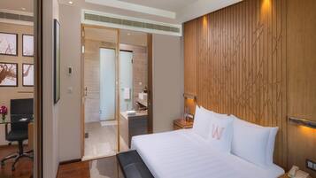Una camera d'albergo moderna con un letto grande, una scrivania e un bagno visibile tramite una porta in vetro.