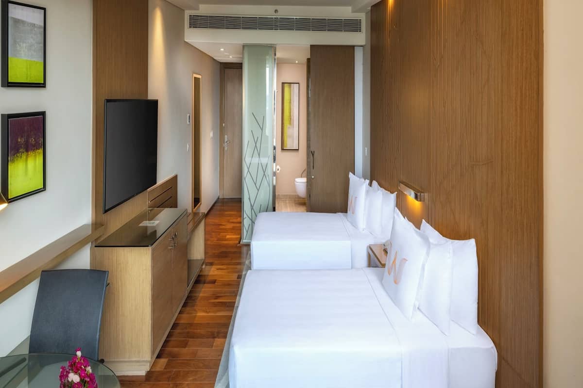 A hotel room with two beds, a TV, a desk, and a bathroom visible through a glass partition.